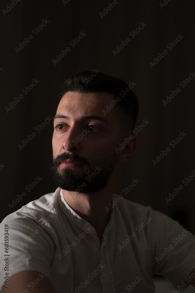 Looking off into the distance with a contemplative, melancholy expression, a young Syrian man poses in a dimly lit room with a single light source illuminating his face. Looking off into the distance with a contemplative, melancholy expression, a young Syrian man poses in a dimly lit room with a single light source illuminating his face.