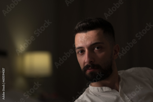 Leaning towards the camera, an attractive, young Syrian man with a black beard and dark eyes gazes mysteriously at the camera. A bedside lamp in the background conveys a domestic, intimate setting.