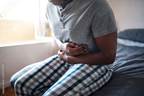 Man, stomach ache and hands holding belly in a bedroom in a home at morning. African male person, sick and indigestion pain in a house on a bed feeling hurt with abdomen cramp and ibs problem photo