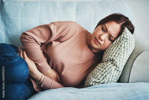 Stomach ache, pain and woman on home sofa with menstrual or period cramps in lounge. Sick, abdomen or colon problem of a female person with hands on tummy for constipation, digestion or virus photo