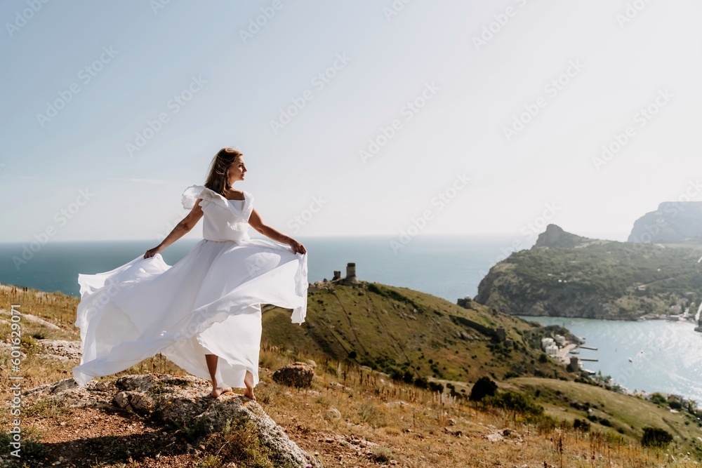 Happy woman in a white dress and hat stands on a rocky cliff above the sea, with the beautiful silhouette of hills in thick fog in the background. Happy woman in a white dress and hat stands on a rocky cliff above the sea, with the beautiful silhouette of hills in thick fog in the background.