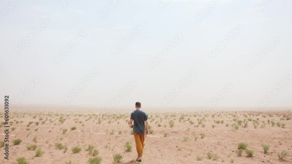 Young man with sunglasses in his hand goes through steppe against backdrop of sandstorm. Back view. Slow motion Young man with sunglasses in his hand goes through steppe against backdrop of sandstorm. Back view. Slow motion