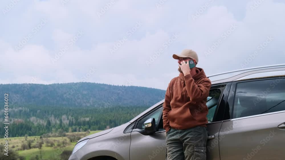Sad man driver stands near the car on a desert country road in mountains. Male traveler gets lost, he is using smartphone calling for help. Road trip trouble.