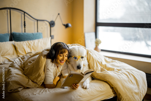 Young woman with her cute dog in eyeglasses reading a book while lying togther under a blanket in bedroom. Spending leisure time, friendship with pet concept photo