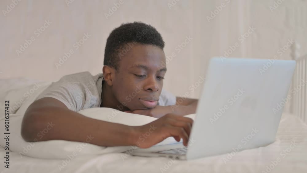 Young African Man Working on Laptop while Lying on Stomach in Bed Young African Man Working on Laptop while Lying on Stomach in Bed