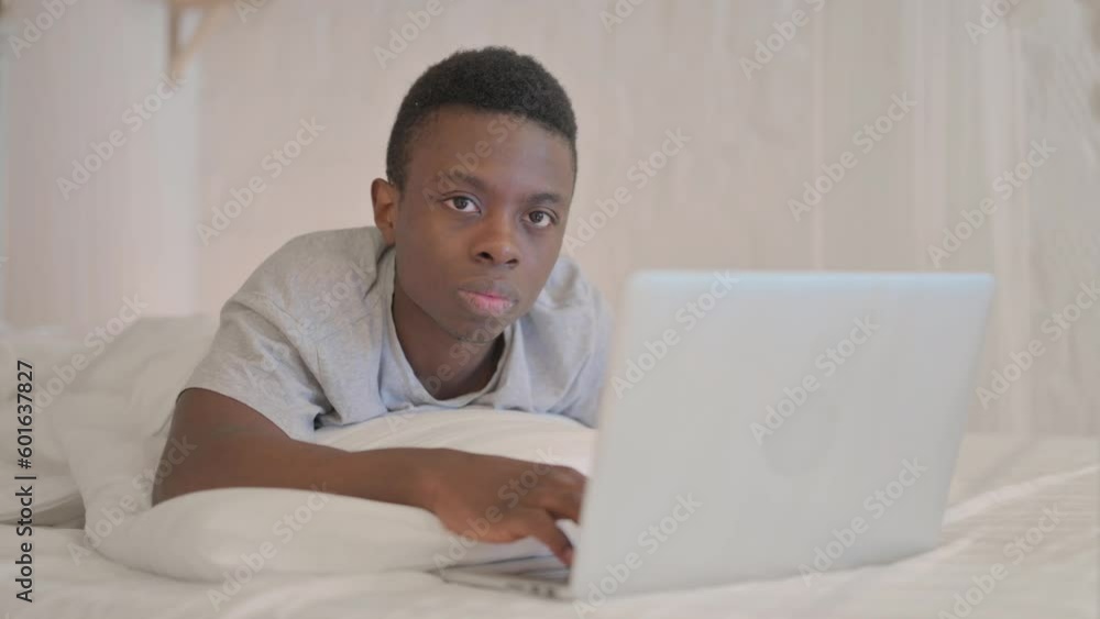 Serious Young African Man Looking in Camera while using Laptop Lying on Stomach Serious Young African Man Looking in Camera while using Laptop Lying on Stomach