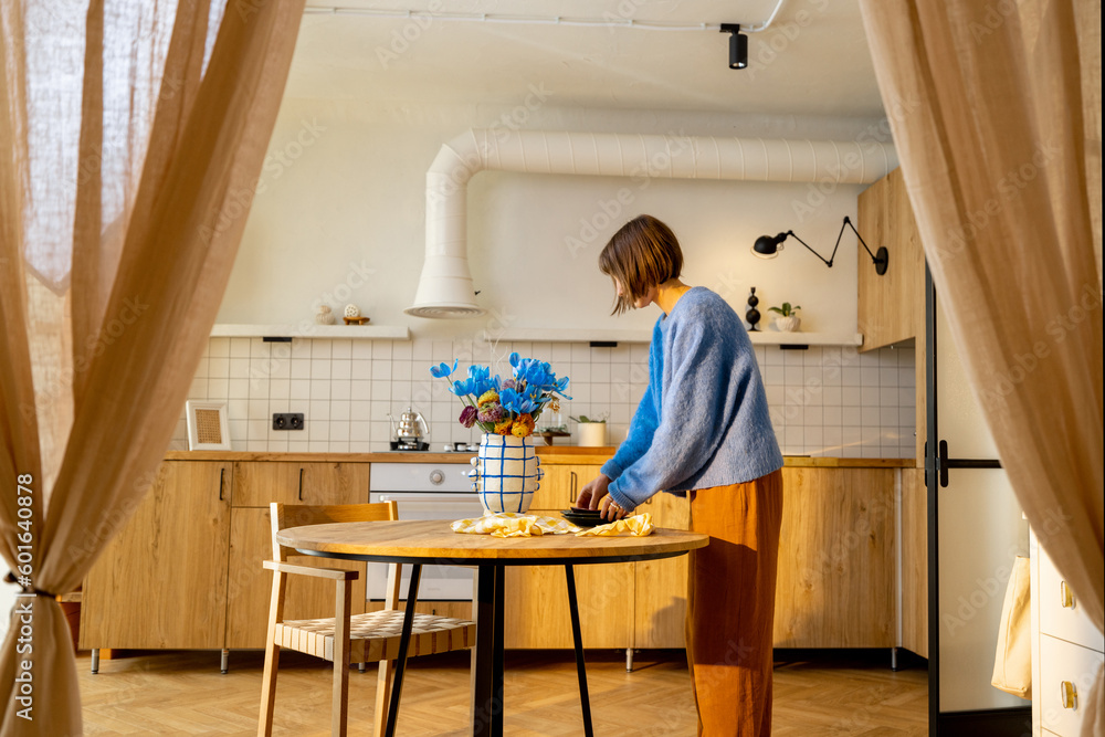 Woman serves a dining table, doing some household chores in stylish studio apartment. Concept of home coziness and lifestyle Woman serves a dining table, doing some household chores in stylish studio apartment. Concept of home coziness and lifestyle