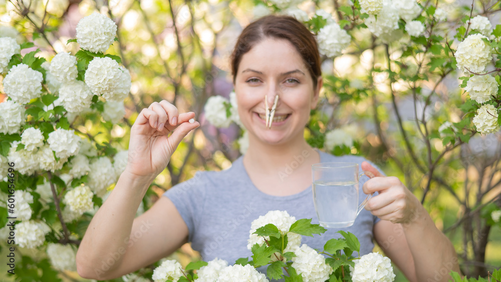 Caucasian woman takes an antihistamine medicine and removes a clothespin from her nose near a flowering tree. Caucasian woman takes an antihistamine medicine and removes a clothespin from her nose near a flowering tree.