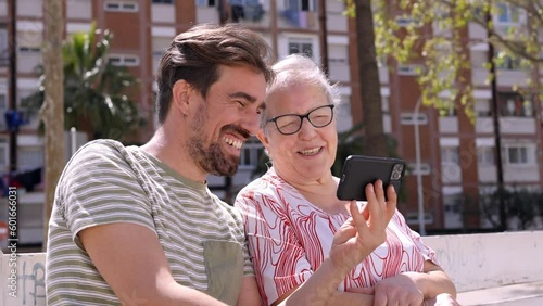 Adult grandson teaches his grandmother how to use smartphone. Sitting outdoors taking a video call together speaking with family. photo