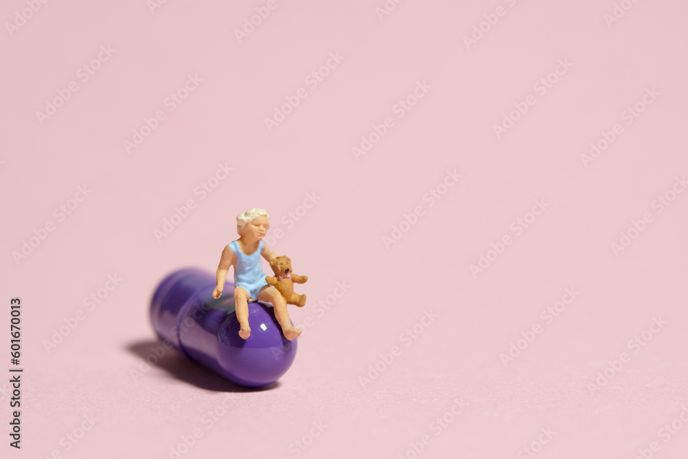 Miniature tiny people toy figure photography. A girl toddler sitting above purple medicine pill with teddy bear. Isolated on pink background Miniature tiny people toy figure photography. A girl toddler sitting above purple medicine pill with teddy bear. Isolated on pink background
