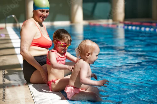 Obraz Happy mother in swimming suit and cap sitting near pool with her little daughters, kids learning how to swim. Concept of sport, healthy and active lifestyle, childhood, fun and training