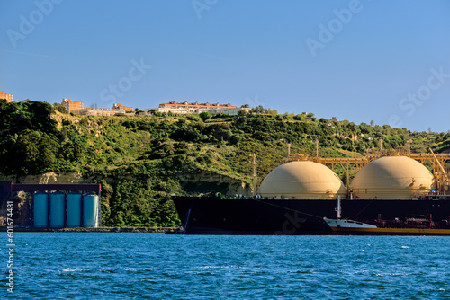 LNG tanker moored by industrial pier and green hill on sunny day