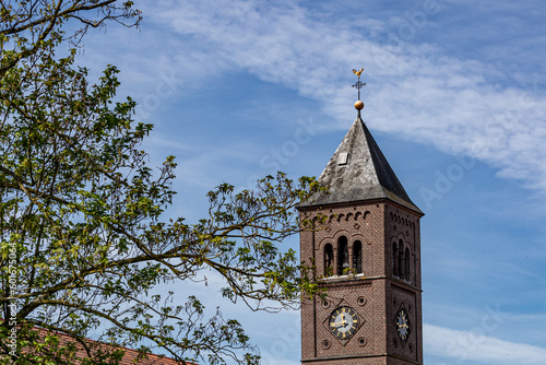 Bell tower of Our Lady Immaculate Conception church in neo-Romanesque style against blue sky, brick walls, clock and green foliage of a tree, spring sunny day in Susteren, South Limburg, Netherlands photo