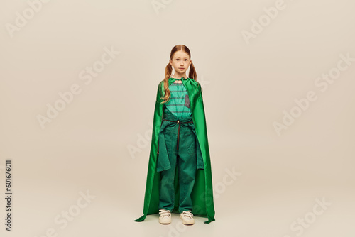 Redhead preteen girl in green superhero costume and cape looking at camera while standing on grey background during global child protection day celebration