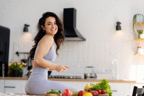 Beautiful smiling fit latin woman holding digital tablet sitting in modern kitchen, cooking fresh salat with fruits and vegetable. Breakfast, diet, healthy lifestyle concept