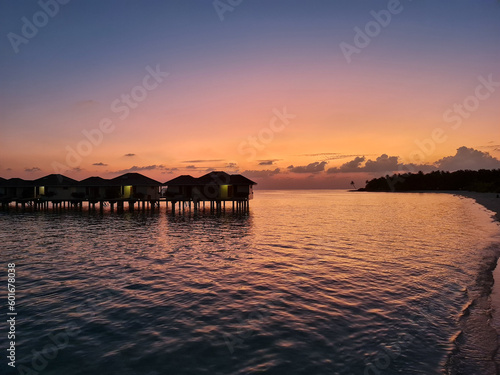 Breathtaking view of the Maldives captured during a sunset over the water with water villas. The water below is calm and reflective, mirroring the colors of the sky above.
