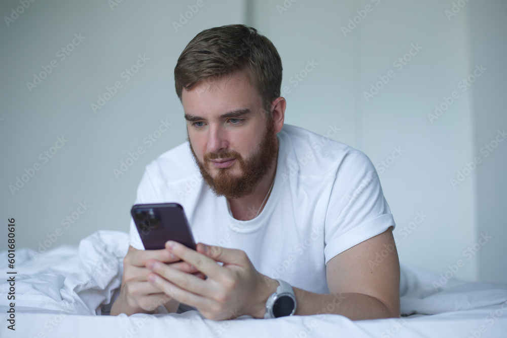 Young bearded man is using cell mobile phone at home in bed in bedroom, looking at screen of smartphone Young bearded man is using cell mobile phone at home in bed in bedroom, looking at screen of smartphone