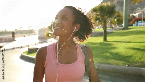 Smiling young African woman in sportswear listening to music on earphones while crossing the street carrying a yoga mat