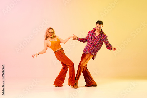 Beautiful, active, emotional couple, man and woman in vintage clothes dancing retro dance against gradient pink yellow background. Concept of retro style, dance, fashion, art, hobby, music, 70s