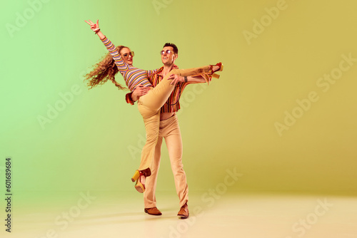 Emotional expressive young man and pretty woman in vintage style outfits dancing retro dance against gradient green yellow background. Concept of retro style, dance, fashion, art, hobby, music, 70s