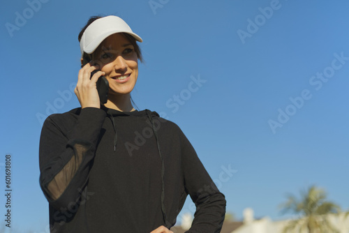 Blond young girl dressed in black sportswear talking on the mobile happily in a park. © Marina