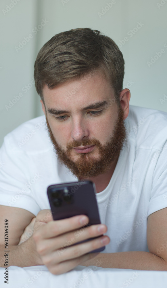 Young happy man wear white t-shirt hold use mobile cell phone browsing internet lying in bed, rest relax spend time in bedroom lounge home in own room house wake up Young happy man wear white t-shirt hold use mobile cell phone browsing internet lying in bed, rest relax spend time in bedroom lounge home in own room house wake up