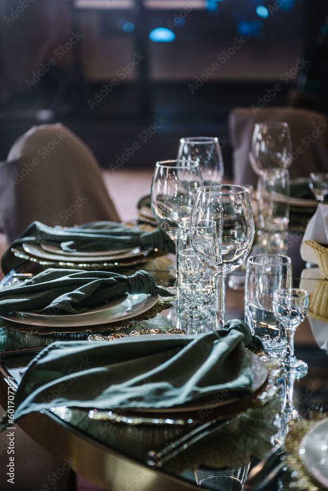 Round served banquet tables for guests, decorated plate, glasses, forks, emerald green napkins. Elegant dinner table. Wedding hall with atmospheric decor. Golden geometric decorative elements decor.