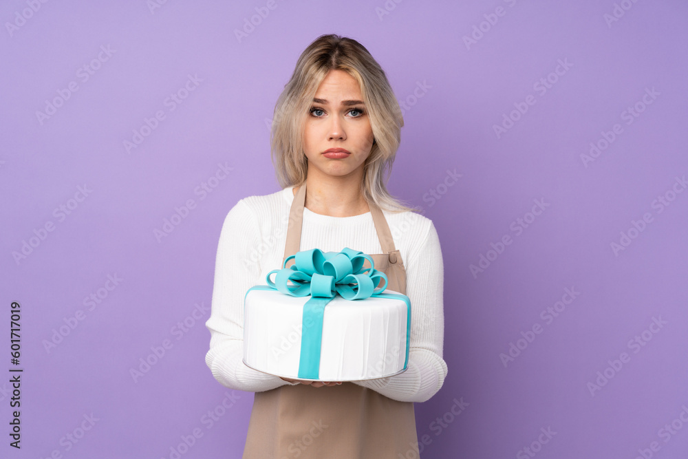 Young pastry chef holding a big cake over isolated purple background sad Young pastry chef holding a big cake over isolated purple background sad