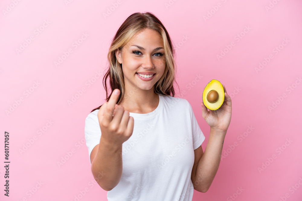 Young Russian woman holding avocado isolated on pink background doing coming gesture Young Russian woman holding avocado isolated on pink background doing coming gesture