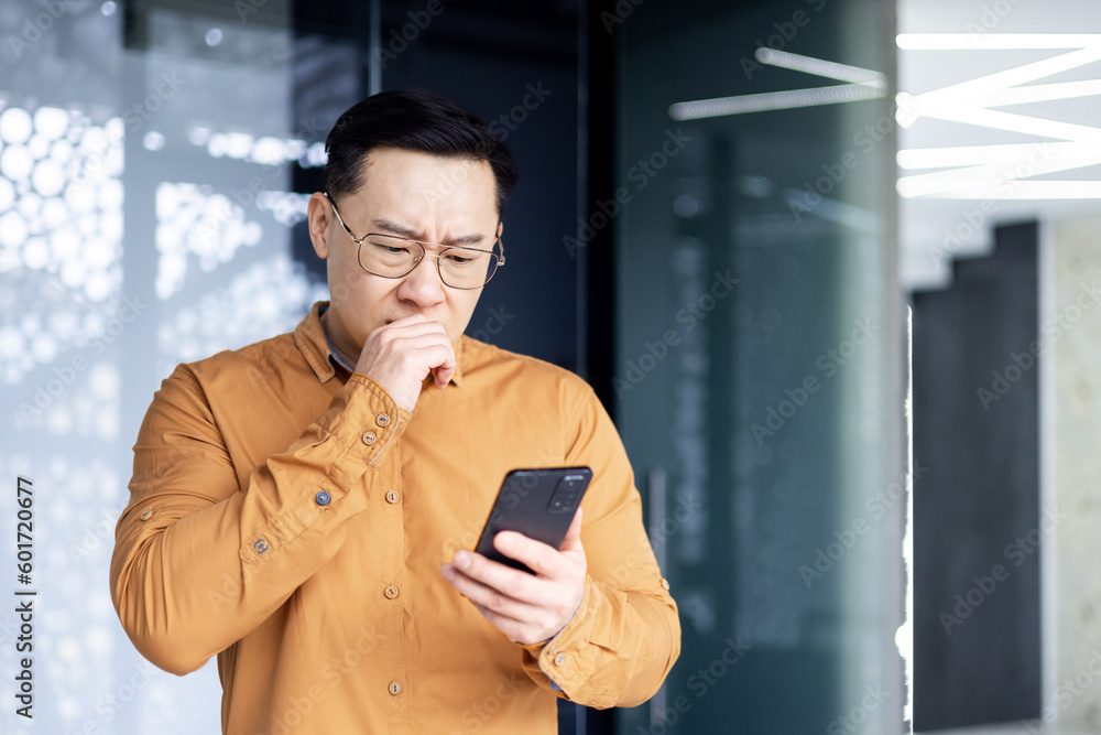 Φωτογραφία Asian businessman inside office building upset and sad reading bad news on mobile phone, business owner investor using smartphone standing near window