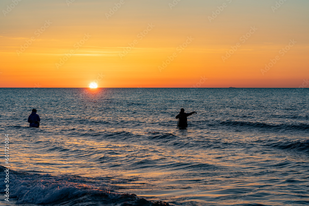 Die Silhouetten von zwei  Anglern in wasserdichten Hosen angelen vor einem traumhaften orangen Sonnenuntergang  in der Ostsee. Sie stehen mitten im Wasser der Ostsee.