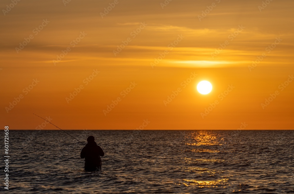 Silhouette eines Angler in wasserdichten Hosen welcher  vor einem traumhaften orangen Sonnenuntergang in der Ostsee angelt / fischt . Er steht mitten im Wasser der Ostsee.