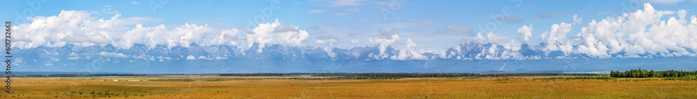 Panorama of foothill Tunka Valley in Buryatia and mountain range of Eastern Sayan Mountains with low cumulus clouds on mountain peaks on sunny day in August. Summer scenic landscape. Nature background