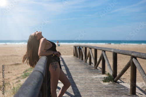 Beautiful young woman with long brown hair in swimming costume and black silk sarong leaning with her head tilted back on the railing of the walkway leading to the beach. The sea in the background.