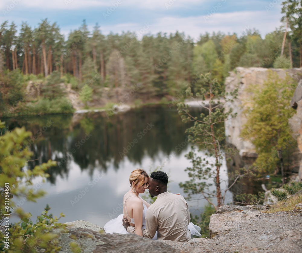 Happy interracial couple newlyweds sits on rock and embraces against beautiful background of lake, forest and canyon.