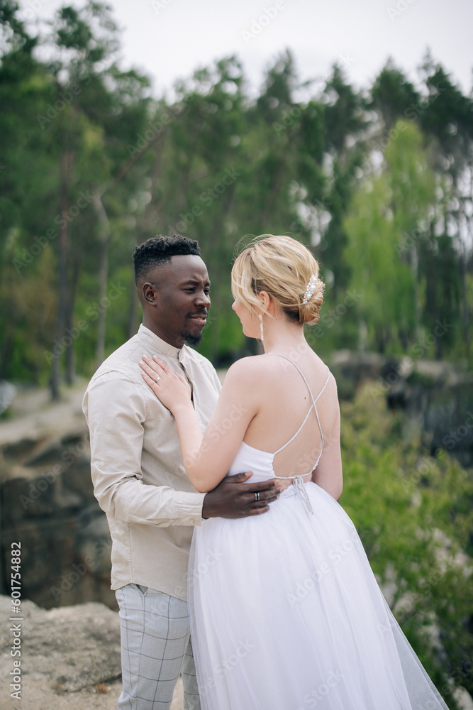 Happy interracial couple newlyweds stands on rock and embraces against background of forest and canyon.