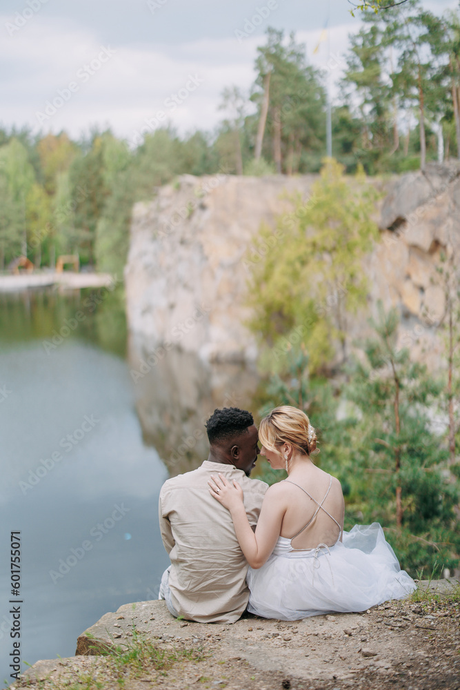 Happy interracial couple newlyweds sits on rock and embraces against beautiful background of lake, forest and canyon.