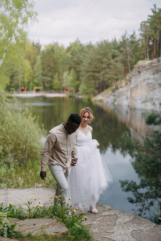 Happy interracial couple newlyweds holds each other hands and walks against background of lake, rocks and forest. Happy interracial couple newlyweds holds each other hands and walks against background of lake, rocks and forest.
