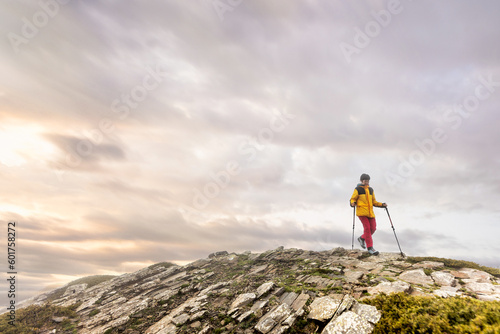 Woman walking with sticks on the mountain at sunset at the top, with colours in the clouds and sun streaks, rocky ground with green bushes, yellow jacket and pink pants.