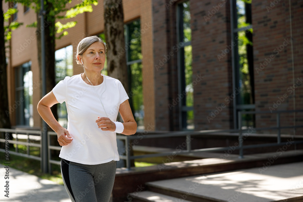 Side view of the attractive smiled Mature Lady running and wearing headphones in the street. Sport and healthy lifestyle concept