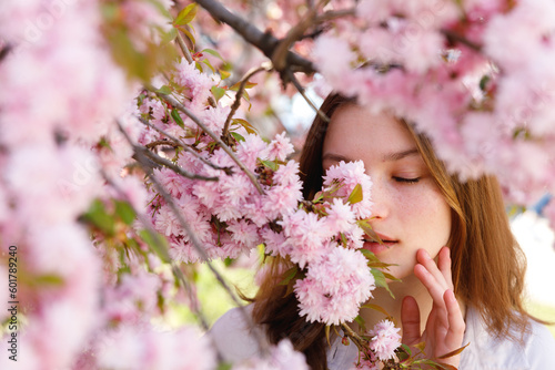 Sensual Beautiful Girl With Brown Hair Covering Half of Face With Branches of Blooming Sakura Tree. Woman Standing in the Park with Closed Eyes. Seasonal Flowering and Femininity Concept. Face