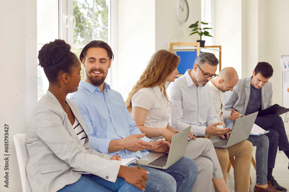 Diverse people communicate with each other during business training in office of business center. Positive people with clipboards and laptops sitting in row on chairs and discussing work. Diverse people communicate with each other during business training in office of business center. Positive people with clipboards and laptops sitting in row on chairs and discussing work.