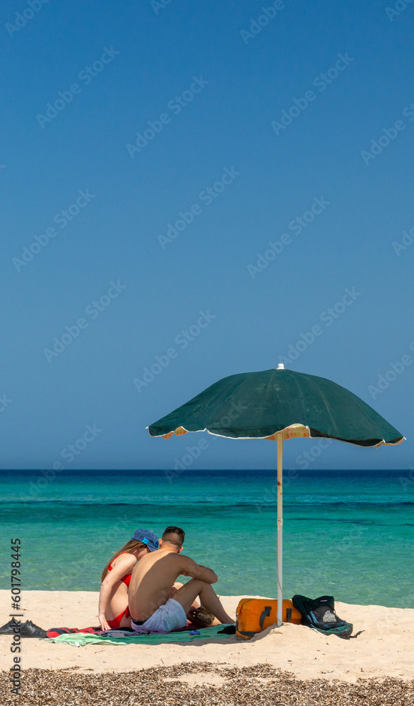 Couple of young persons on the beach with umbrella on golden sand with blue sky and crystal turquoise sea.
