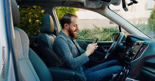 Bearded young man sitting in car and typing online message or reading news on cellphone. Male sitting in modern vehicle and using on smartphone. © serg