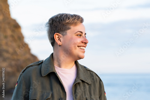 Cheerful Gaze: Non-Binary Person Smiling by the Seaside in Pink Shirt and Green Military Jacket