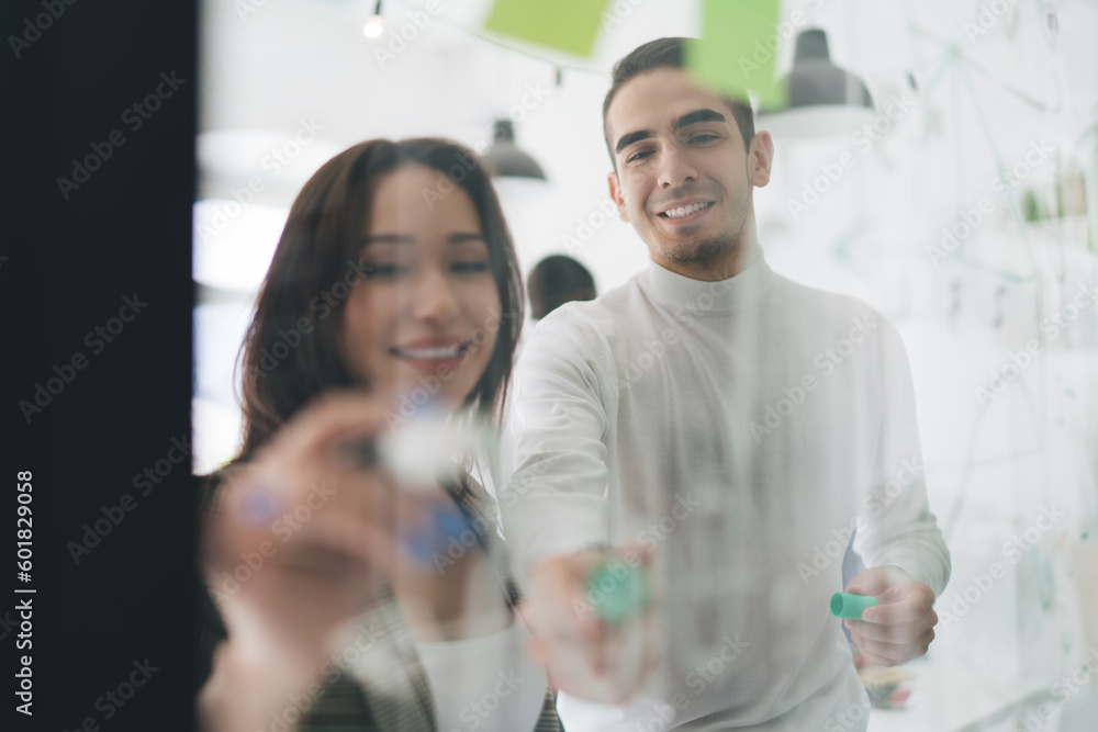 Obraz premium Cheerful young colleagues standing and working on project at glass wall in office