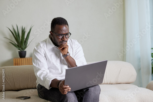 Handsome Black African American Man Working on Laptop Computer while Sitting on a Sofa in Cozy Living Room. Freelancer Working From Home. Browsing Internet, Using Social Networks, Having Fun in Flat.