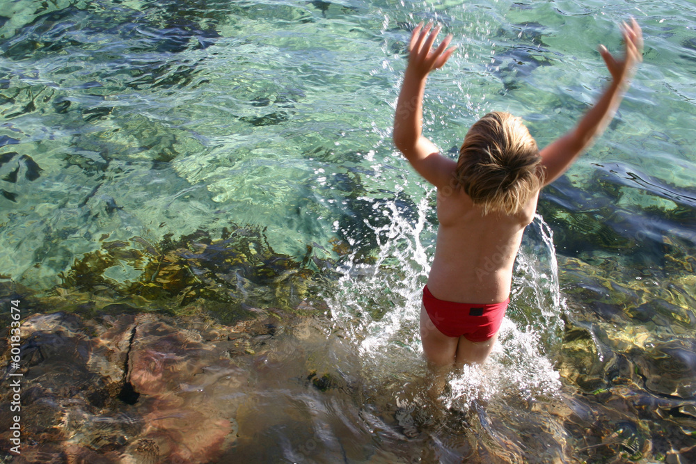 Fototapeta premium Boy splashes water overhead from an ocean rockpool. Some motion blur in the arms and hands.