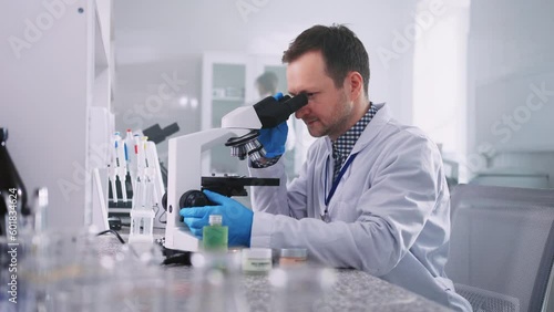Footage of young Caucasian man in white labcoat looking through microscope at laboratory. Shot of man studying microbiological substance in microscope and using lab jar. Scientist at work. Indoors