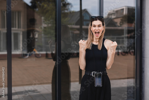 Young excited confident proud business woman winner wearing black outfit standing on street, raising hands, feeling power, motivation, energy, celebrating career financial success in big city outdoors